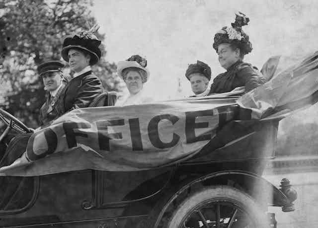 A black and white photo shows four middle aged women with fancy clothing and hats riding in an old car that says 'officer' on the side.