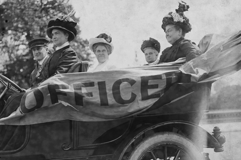 A black and white photo shows four middle aged women with fancy clothing and hats riding in an old car that says 'officer' on the side.