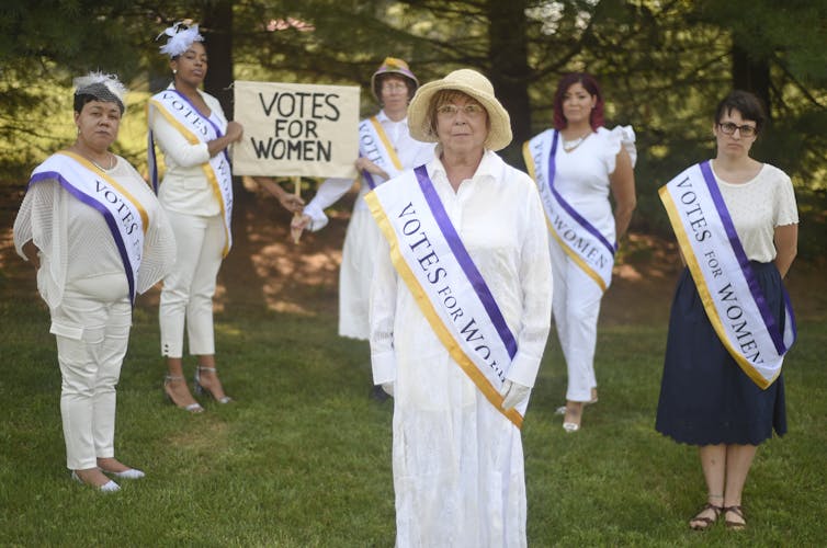 A middle aged white, blonde woman wears a straw hat, white clothing and a sash that says 'Votes for women.' Five other women, also in white and with the same sashes, stand in the foreground. Two hold a sign that says 'Votes for Women.'