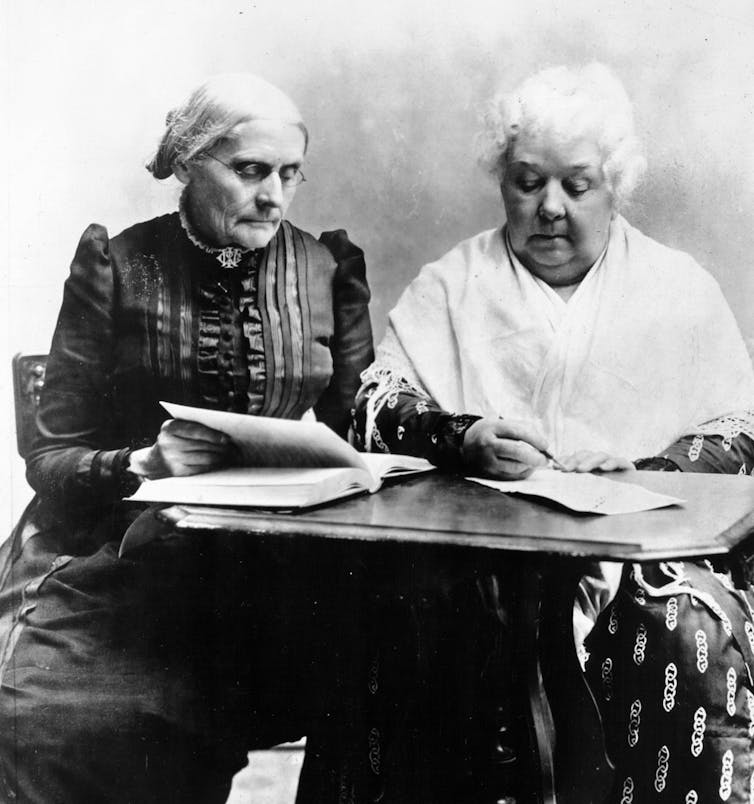 A black and white photo shows two older women in old fashioned clothing seated at a table, looking at some papers.