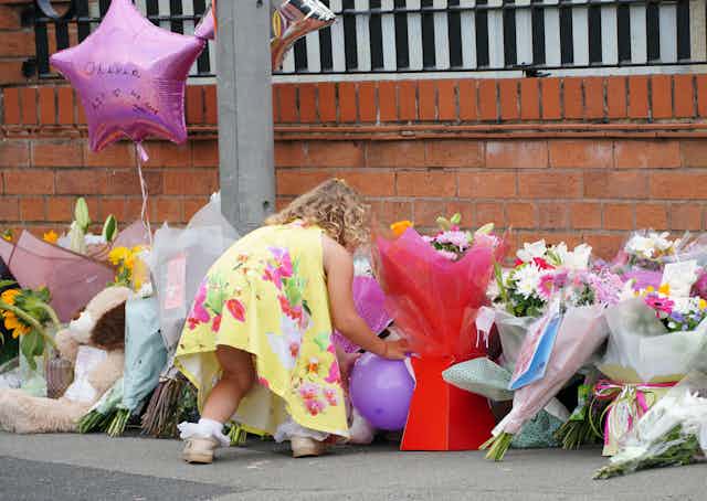 A young girl in a yellow dress places a balloon among a pile of bouquets and stuffed animals paying tribute to Olivia Pratt Korbel
