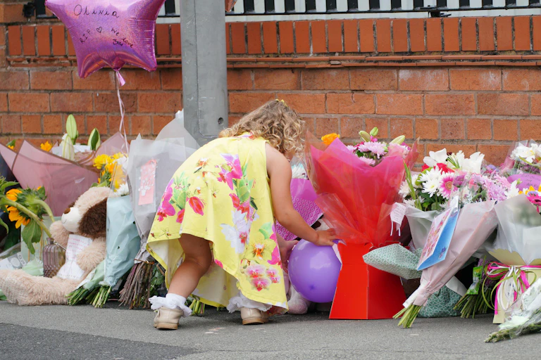 A young girl in a yellow dress places a balloon among a pile of bouquets and stuffed animals paying tribute to Olivia Pratt Korbel