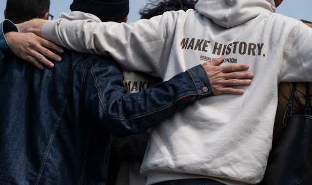 Pro-union workers stand with their backs to the camera with arms linked. A slogan on the back of a hoodie reads 'Make History. Amazon Union.