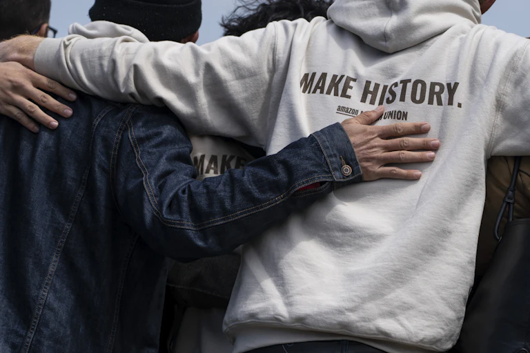 Pro-union workers stand with their backs to the camera with arms linked. A slogan on the back of a hoodie reads 'Make History. Amazon Union.