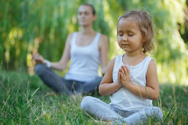 A young girl, sitting in a grassy field, meditates with her mother, who sits nearby, also meditating.