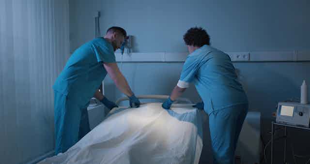 Dos celadores tapan con una sábana blanca el cuerpo de un paciente hospitalario fallecido.