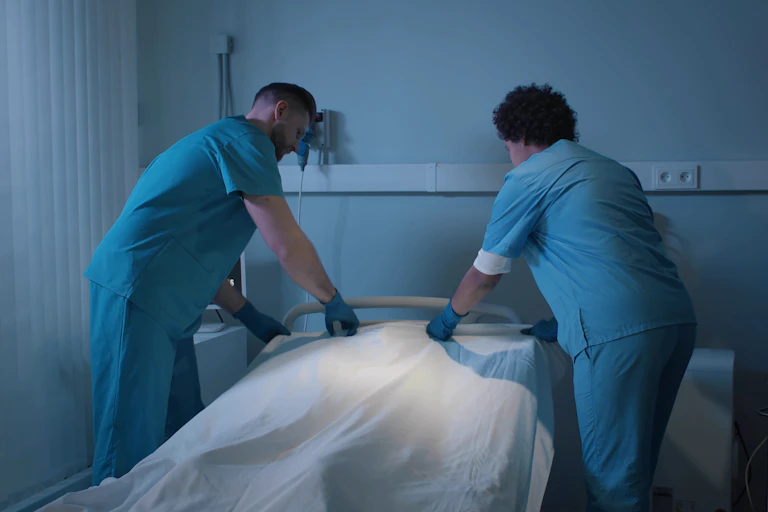 Dos celadores tapan con una sábana blanca el cuerpo de un paciente hospitalario fallecido.