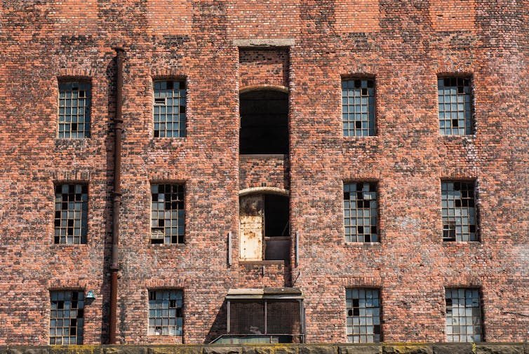 Front facade of a derelict industrial red brick warehouse, with many broken windows.