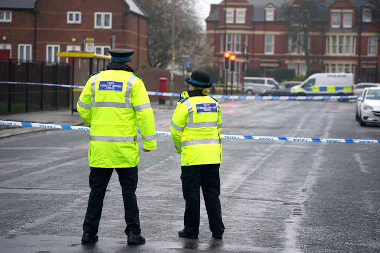 Two community support officers in high-vis jackets stand in a car park next to police tape