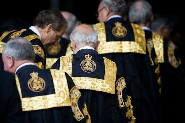 Old white men in black robes with gold trim are seen walking from behind.