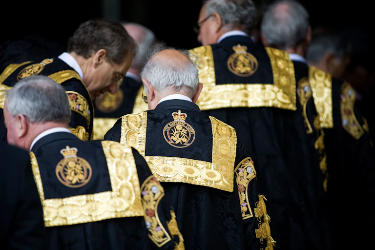 Old white men in black robes with gold trim are seen walking from behind.