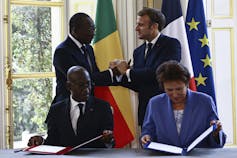 Presidents of France and Benin shake hands. In front of the two other people sit at a desk holding documents.