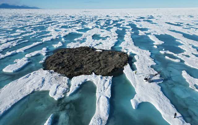 A helicopter sits on ice next to what appears to be a gravel-covered island, but is actually an iceberg. People are standing on its surface.