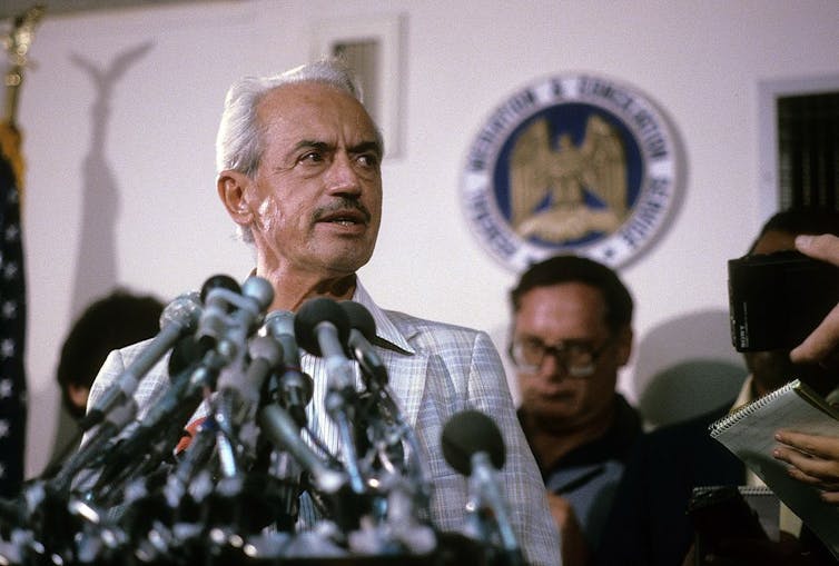 Man with mustache speaks in front of microphones.