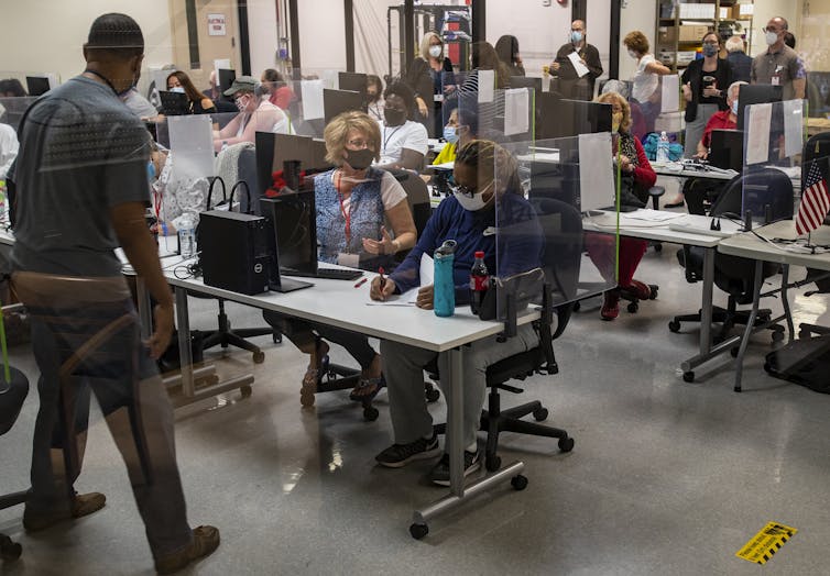 A Black and a white woman sit side by side at a table with a desktop computer in a crowded room.