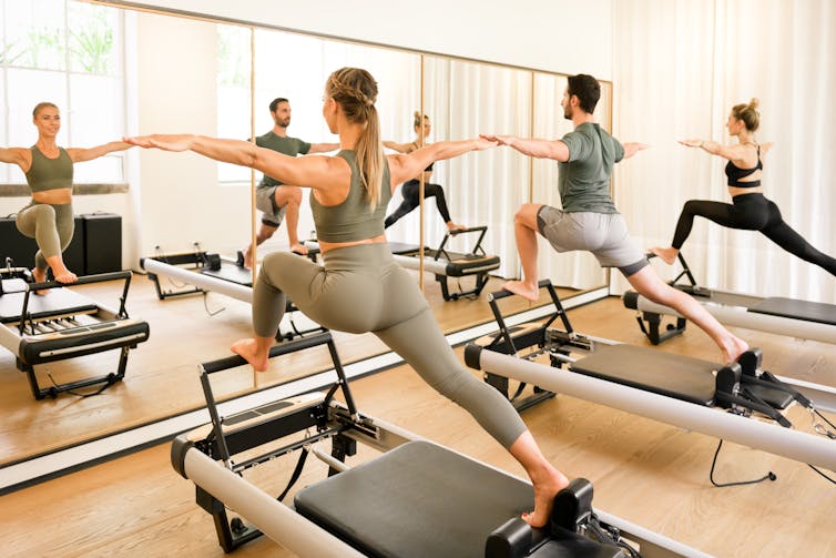 Two women and one man perform a pilates move using a pilates reformer. They are lunging forward with one leg and holding their arms out.