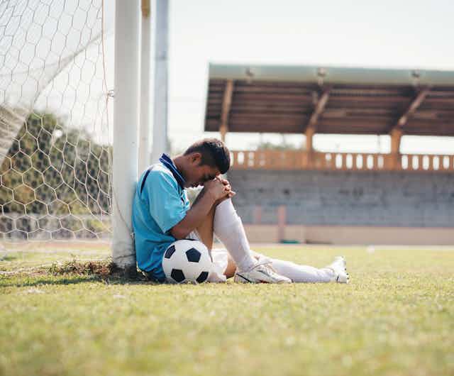 Sad boy with ball sat on football pitch