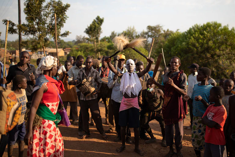 A crowd in a village, at the centre of attention a young man holding up two sticks trimmed with animal hair. His face has white clay on it. People gather around him, some with sticks or drums.