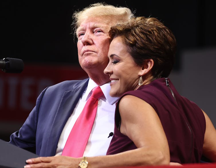 A white man dressed in navy blue suit with a white shirt and red tie hugs a smiling woman on stage.