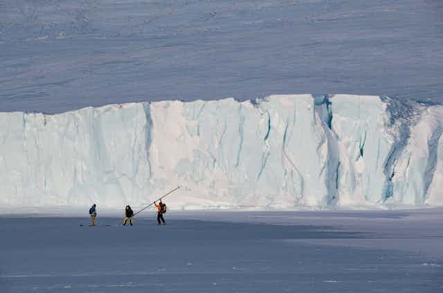 Three figures in the distance raising a flagpole in front of a glacier