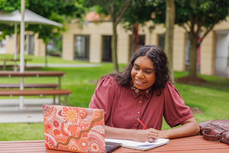A smiling First Nations woman sits at a laptop with an Aboriginal art design, writing in a notebook