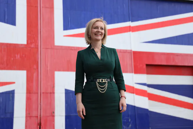 Liz Truss yang mengenakan gaun hijau berdiri dan tersenyum di depan dinding yang dicat sebagai union jack