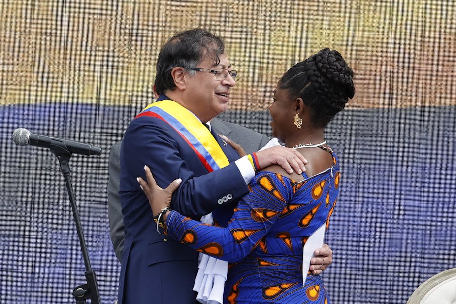 A man wearing specs, left, and a woman with braided hair smile as they embrace next to a microphone.