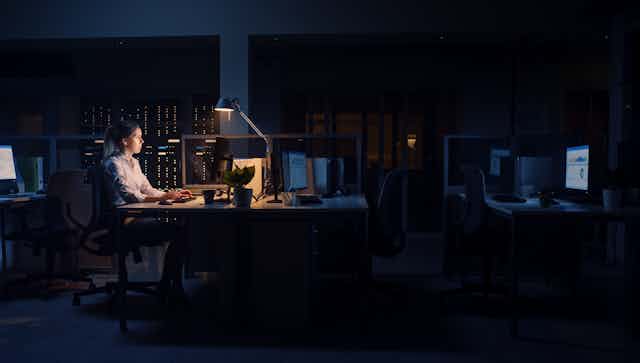 A woman working at her desk in a dark office, with just her lamp illuminating her workspace
