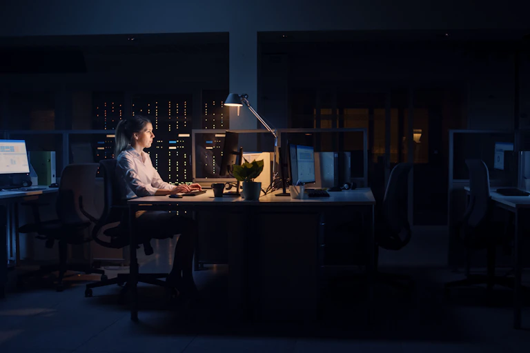 A woman working at her desk in a dark office, with just her lamp illuminating her workspace