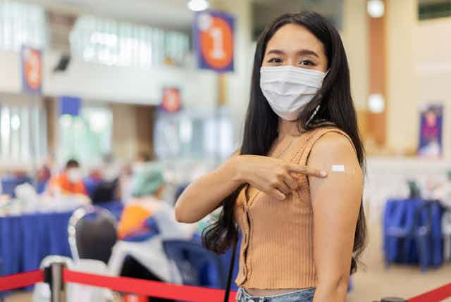 A young woman wearing a mask points to a plaster on her upper arm.
