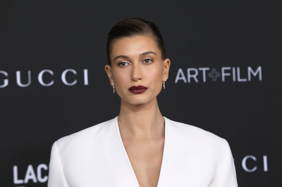 A picture of model Hailey Bieber at a fashion event. She is wearing dark red lipstick and a white suit.