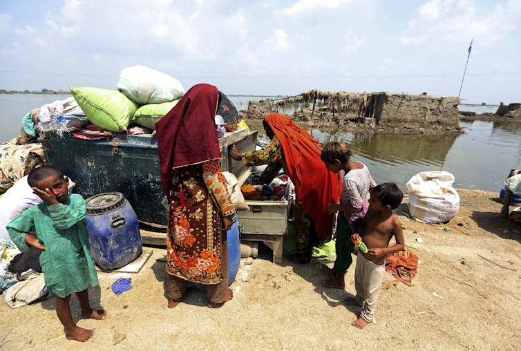 women stand on a sandy beach with household belongings piled high on a cart