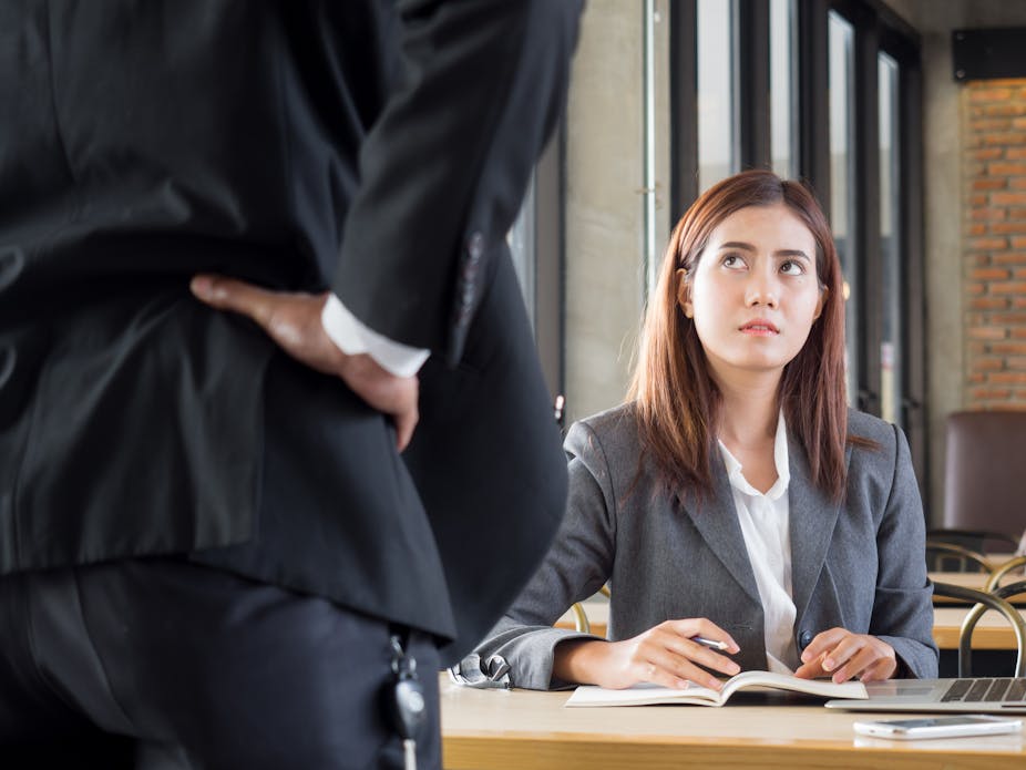 A young woman sits at her desk looking worried, while in the foreground her boss has his hands on his hips