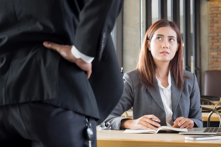 A young woman sits at her desk looking worried, while in the foreground her boss has his hands on his hips