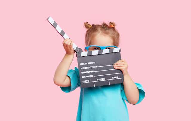 A girl wearing blue glasses holds a clapperboard in front of her face.