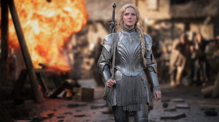 A woman in battle armour walks away from a fire.