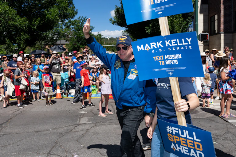 A man wearing a baseball hat and blue jacket is waving to a crowd of people gathered to watch a parade.