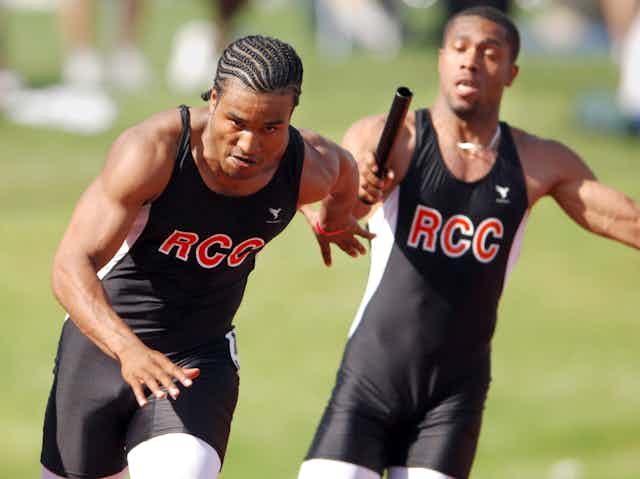 One runner from a community college track and field team hands a baton to another runner.