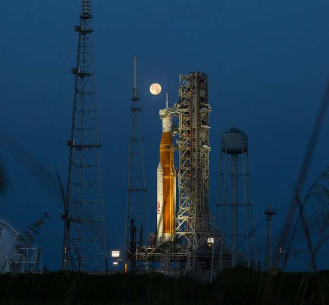 An orange and white rocket at night at the launch pad, with a full moon shining on it