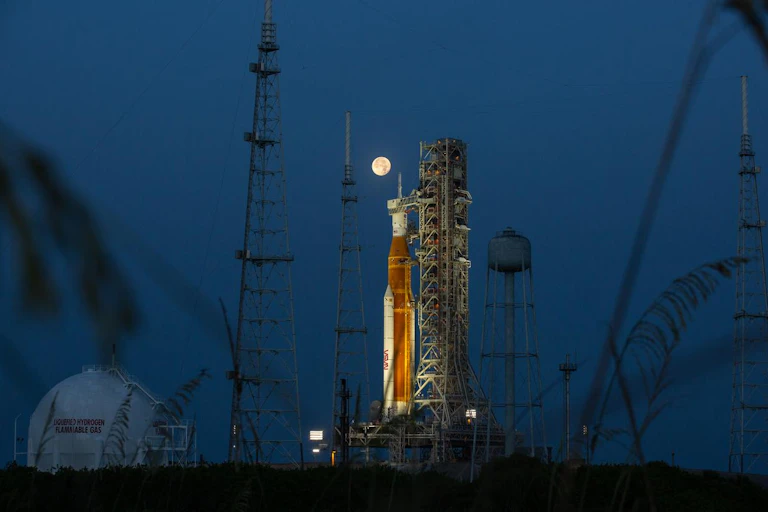 An orange and white rocket at night at the launch pad, with a full moon shining on it