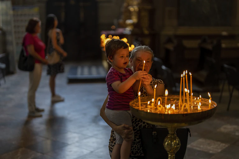An elderly woman helps a child light candles in a church.