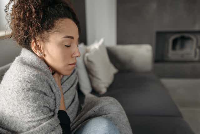 Woman wrapped in blanket, hand at throat, eyes shut, sick on sofa at home