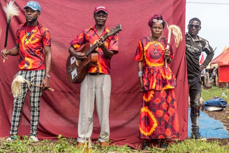 Four people against a backdrop of dark pink sheet. They are colourfully dressed in traditional attire, a man holding a guitar.