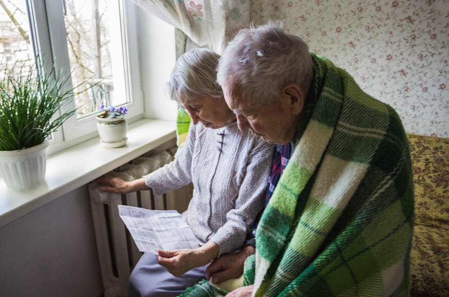 An elderly couple looks down at an energy bill, the man is wrapped in a green plaid blanket while the wife rests her hand on the radiator