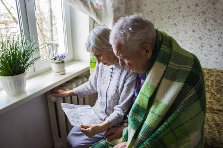 An elderly couple looks down at an energy bill, the man is wrapped in a green plaid blanket while the wife rests her hand on the radiator