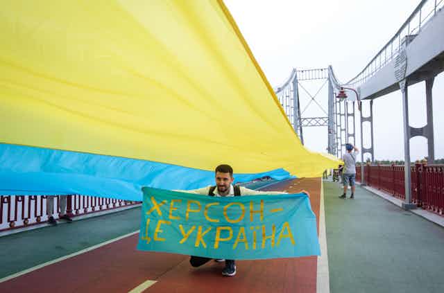 A man holds a banner reading 'Kherson is Ukraine' against the background of a large Ukrainian flag on a bridge over the Dnipro River in Kyiv, August 2022.