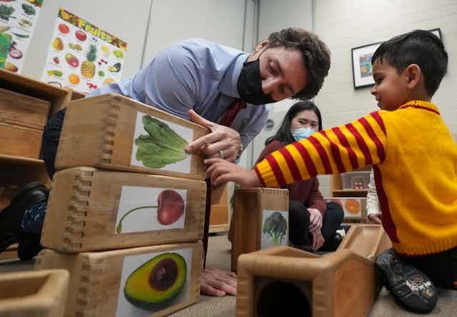 On voit un homme masqué, Justin Trudeau, montrer du doigt une feuille de laitue sur un bloc, tandis qu'un garçon dans une garderie sourit et écoute.