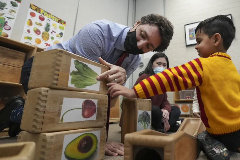 On voit un homme masqué, Justin Trudeau, montrer du doigt une feuille de laitue sur un bloc, tandis qu'un garçon dans une garderie sourit et écoute.