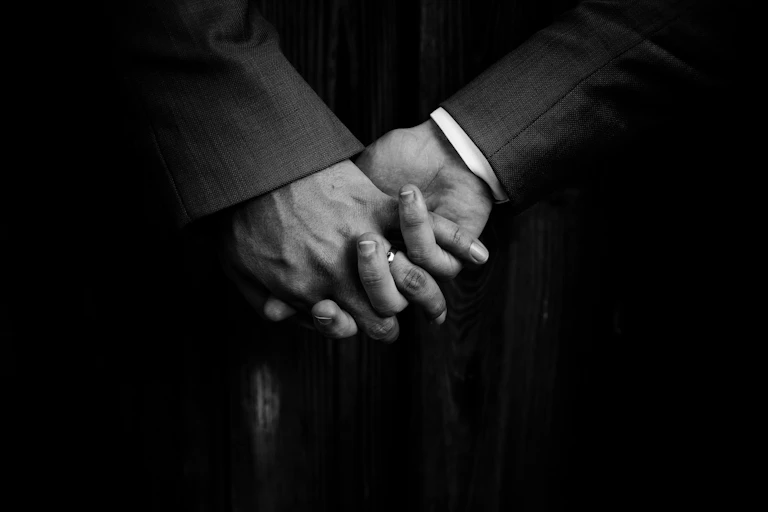 A black and white photograph shows the clasped hands of two people in suit jackets. One has a visible wedding ring.