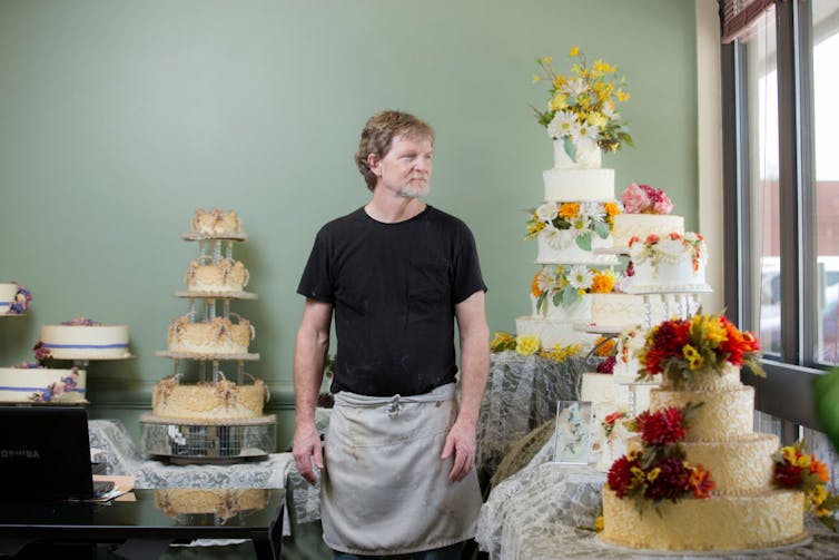 A man in a black shirt and gray apron stands among tiered wedding cakes in a green room.